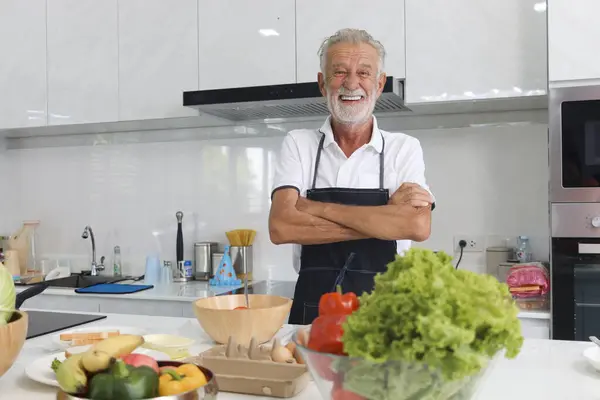 Portrait of happy elderly man with apron standing with arms crosse at kitchen with colorful fresh vegetables, fruits,ingredients. Senior man cooking healthy food at home, mature grandfather make meal.