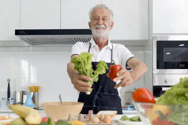 Elderly man holding fresh sweet red bell pepper and green leaf vegetable for making salad, senior man cooking healthy food with organic ingredients at home, mature grandfather making meal at kitchen.