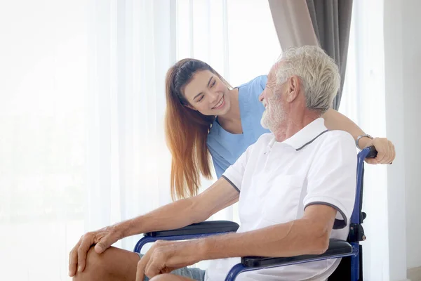 Beautiful female nurse taking care of senior patient in wheelchair at home, disabled elderly man sitting in wheelchair, young woman doctor helping of mature grandfather, nursing medical health care.