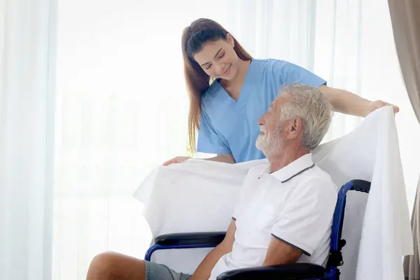Beautiful female nurse takes care of senior patient in wheelchair at home, disabled elderly man sits in wheelchair while woman doctor covers blanket to mature grandfather, nursing medical health care.
