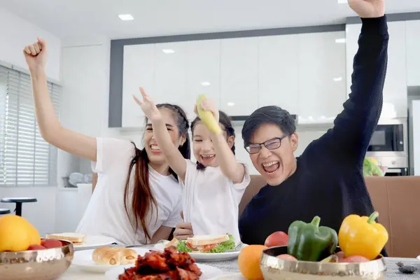 Happy family has meal in dining room. Mother father kid daughter sit at dining table, have fun during breakfast or lunch. Cheerful family raising hand up, parent and child enjoy eating food together.