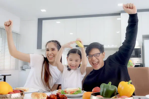 Happy family has meal in dining room. Mother father kid daughter sit at dining table, have fun during breakfast or lunch. Cheerful family raising hand up, parent and child enjoy eating food together.