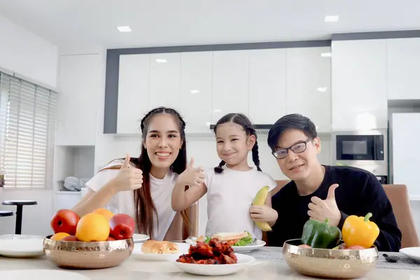 Happy family has meal in dining room. Mother father kid daughter sit at dining table, have fun during breakfast or lunch. Cheerful family giving thumbs up, parent and child enjoy eating food together.