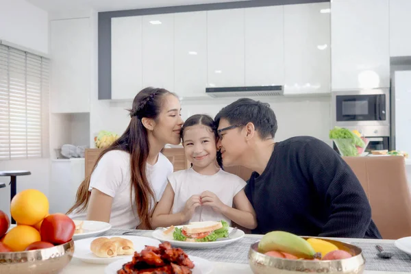 Happy family has meal in dining room. Mother father kid daughter sit at dining table, have fun during breakfast, parent kiss girl child who make heart shape by hands. Family enjoy eating food together