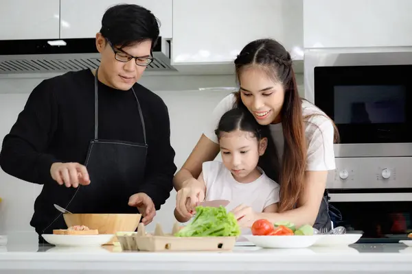 Happy family cooking at kitchen, father mother and cute daughter girl having fun during making meal at home. Parents and kid child enjoy cooking food, spending time together. Family love and bonding.