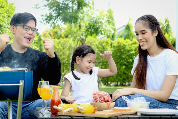 Joyful parent and kid child make barbecue at picnic party, have fun together in summer green garden, father mother and daughter girl enjoy eating and drinking at backyard, happy family love bonding.