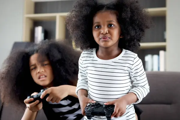 Joyful African sister girl with black curly hair enjoy play video game at home, two children hold console joystick controller to play game, happy kid siblings be friend and spend free time together.