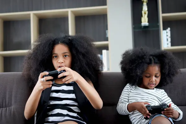 Joyful African sister girl with black curly hair enjoy play video game at home, two children hold console joystick controller to play game, happy kid siblings be friend and spend free time together.