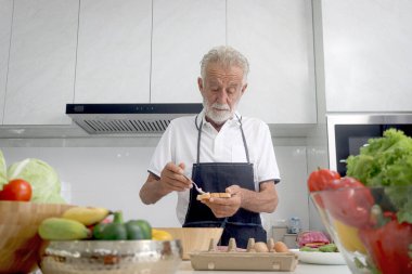 Mutfak tezgahında önlük giyip renkli taze sebzeler ve meyvelerle aç yaşlı bir adam. Sandviç yapmak için ekmek tutan kıdemli adam, olgun büyükbaba evde tek başına yemek pişirmekten hoşlanır..