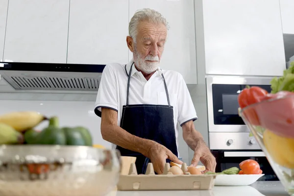 Hungry elderly man wearing apron at kitchen counter with colorful fresh vegetables and fruits. Senior man holding bread for making sandwich, mature grandfather enjoys cooking meal by himself at home.