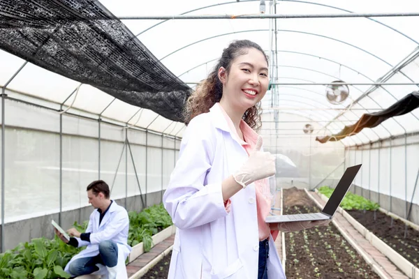 Happy smiling beautiful Asian botanist scientist woman in lab coat hold laptop computer and give thumb up, biological researcher works on experimental plant plots in greenhouses. Agricultural Science
