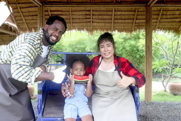 Happy farmer family work together in agriculture, mother and father gardener spend time with daughter kid, African Asian mixed race curly hair girl enjoy eating watermelon product from their own farm.