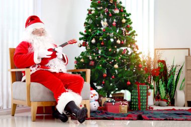 Kırmızı kostümlü Noel Baba Ukulele çalıyor ve oturma odasında dekoratif Noel ağacının önünde Noel şarkıları söylüyor. Mutlu büyükbaba beyaz sakallı Noel Baba kış tatilini kutluyor. Mutlu Noeller.