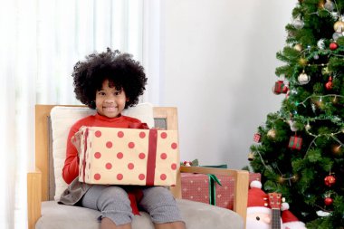Mutlu gülümseyen kıvırcık saçlı Afrikalı kız çocuğunun portresi, arka planda güzel dekoratif Noel ağacıyla birlikte Noel hediyesi kutusunu tutuyor. Çocuk kış tatilini kutluyor
