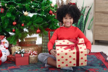 Süslü Noel ağacının altında otururken elinde hediye kutusu olan kıvırcık saçlı mutlu Afrikalı kız çocuğunun portresi. Çocuk kış tatilini kutlar. Mutlu Noeller, mutlu çocukluk