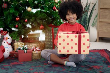 Süslü Noel ağacının altında otururken elinde hediye kutusu olan kıvırcık saçlı mutlu Afrikalı kız çocuğunun portresi. Çocuk kış tatilini kutlar. Mutlu Noeller, mutlu çocukluk