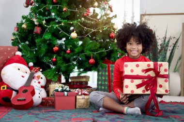 Süslü Noel ağacının altında otururken elinde hediye kutusu olan kıvırcık saçlı mutlu Afrikalı kız çocuğunun portresi. Çocuk kış tatilini kutlar. Mutlu Noeller, mutlu çocukluk