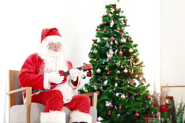 Santa Claus in red costume playing Ukulele and singing Christmas songs front of decorative Christmas tree in living room. Happy grandpa white beard Santa celebrating winter holiday. Merry Christmas
