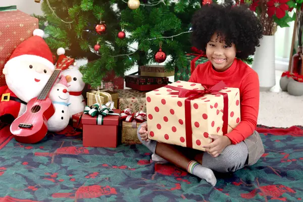 Süslü Noel ağacının altında otururken elinde hediye kutusu olan kıvırcık saçlı mutlu Afrikalı kız çocuğunun portresi. Çocuk kış tatilini kutlar. Mutlu Noeller, mutlu çocukluk