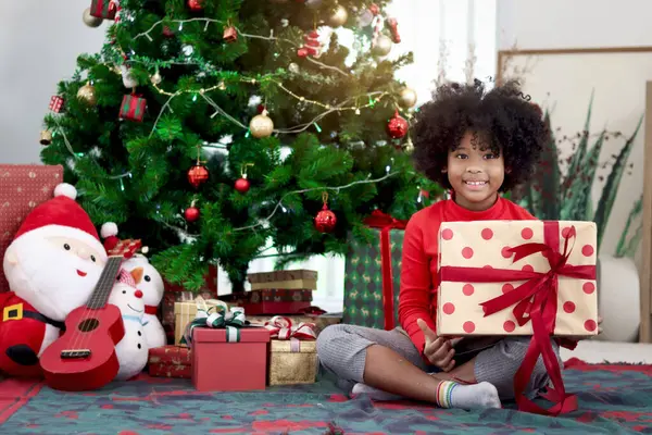 Süslü Noel ağacının altında otururken elinde hediye kutusu olan kıvırcık saçlı mutlu Afrikalı kız çocuğunun portresi. Çocuk kış tatilini kutlar. Mutlu Noeller, mutlu çocukluk