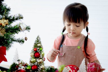 Küçük tatlı Asyalı kız çocuğu beyaz oturma odasında güzel bir Noel ağacını süsleyip süslüyor. Kış tatilini kutlayan çocuğa konsantre ol. Mutlu Noeller ve mutlu yıllar. Mutlu çocukluk