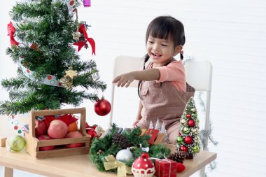 Küçük tatlı Asyalı kız çocuğu beyaz oturma odasında güzel bir Noel ağacını süsleyip süslüyor. Kış tatilini kutlayan çocuğa konsantre ol. Mutlu Noeller ve mutlu yıllar. Mutlu çocukluk