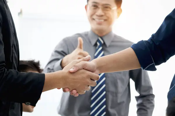 Businesspeople shaking hands after meeting with blurred background of businessman giving thumb up to admire compliment, handshake reached agreement on negotiation deal, successful unity partnership.