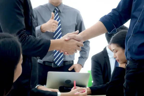 Businesspeople shaking hands after meeting with blurred background of businessman giving thumb up to admire compliment, handshake reached agreement on negotiation deal, successful unity partnership.