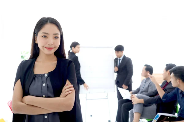 Portrait of Asian beautiful businesswoman standing with arms crossed with blurred background of businesspeople group at conference. Happy smiling woman at office meeting. female officer at workplace.