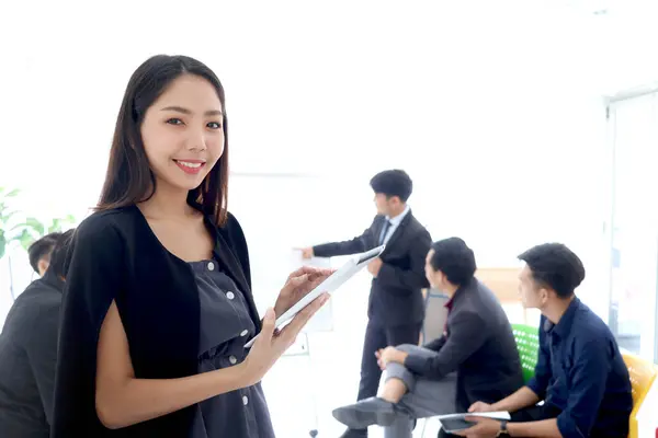Portrait of Asian beautiful businesswoman holding digital tablet with blurred background of businesspeople group at conference. Happy smiling woman at office meeting. female officer at workplace.