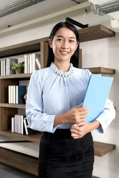 Portrait of happy smiling beautiful Asian woman holding file paper document, standing inside office building, female officer staff in casual uniform, confident businesswoman at workplace.