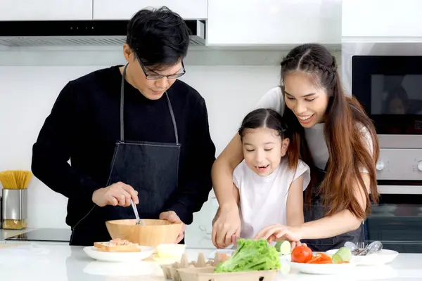 Happy family cooking at kitchen, father mother and cute daughter girl having fun during making meal at home. Parents and kid child enjoy cooking food, spending time together. Family love and bonding.