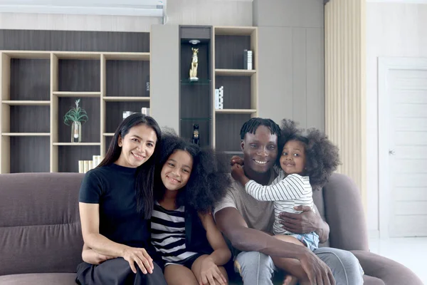 Happy family love bonding, African father Asian mother and two daughter girls with curly hair enjoy spending time together in living room at home, kids and parents hugging each other sitting on sofa.