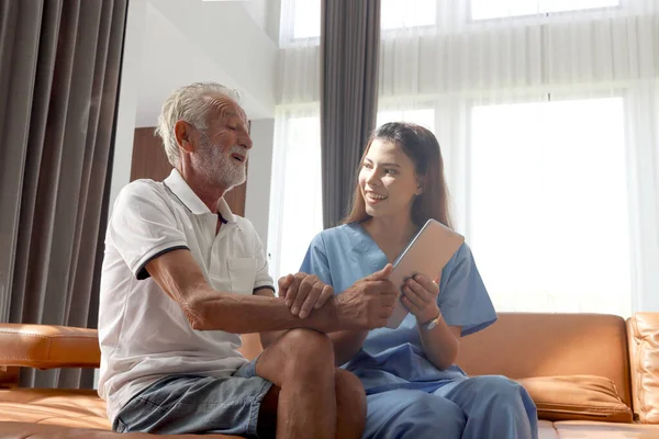 Female nurse takes care of senior patient and teaches him to use digital tablet at living room house, elderly mature grandfather learns to online internet and high-tech, nursing medical health care.