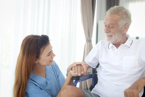 Happy senior patient in wheelchair with beautiful female nurse taking care of him at home, disabled elderly man sits in wheelchair, woman doctor helping mature grandfather, nursing medical health care