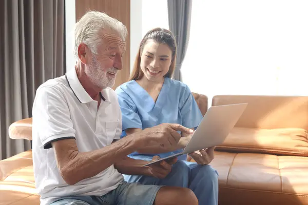 Female nurse takes care of senior patient and teaches him to use laptop computer at living room house, elderly mature grandfather learns to online internet and high-tech, nursing medical health care.