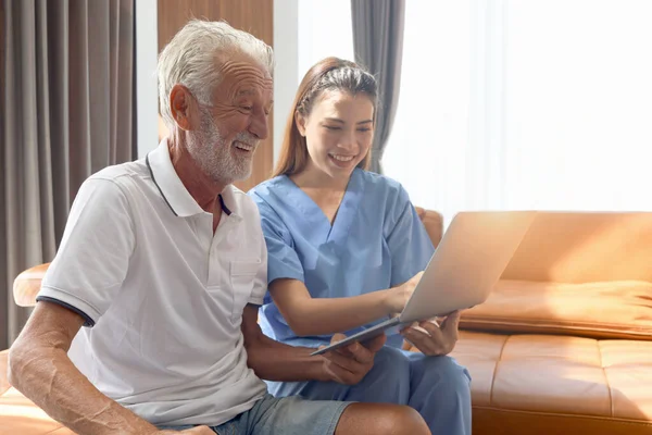 Female nurse takes care of senior patient and teaches him to use laptop computer at living room house, elderly mature grandfather learns to online internet and high-tech, nursing medical health care.