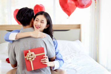 Mutlu Asyalı çift yıldönümünü birlikte kutluyor. Sevgilisi, sevgilisine sürpriz bir hediye kutusu verdikten sonra sevgilisine sarılıyor. Sevgililer gününü güzel dekore edilmiş yatak odasında kutluyor..