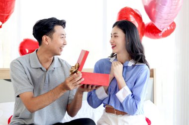 Mutlu Asyalı çift birlikte aşklarını kutluyor, erkek arkadaş sürpriz sevgiliye kırmızı hediye kutusu veriyor, romantik aşık sevgililer gününü güzel dekore edilmiş yatak odasında kutluyor.