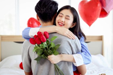 Mutlu Asyalı çift yıldönümlerini birlikte kutluyor. Sevgilisi, sevgililer gününde güzel dekore edilmiş yatak odasında ona sürpriz yapmak için kırmızı gül buketi verdikten sonra sevgilisine sarılıyor..