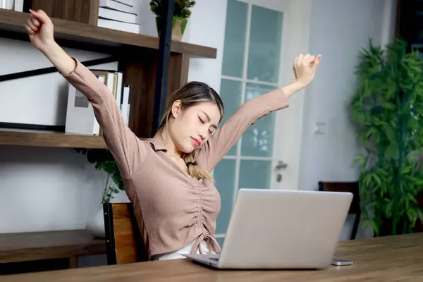 Tired beautiful Asian woman worker relieve physical tension after work hard at home, exhausted female worker feels sleepy, reaching hand and stretching body at working desk.