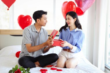 Mutlu Asyalı çift birlikte aşklarını kutluyor, erkek arkadaş sürpriz sevgiliye kırmızı hediye kutusu veriyor, romantik aşık sevgililer gününü güzel dekore edilmiş yatak odasında kutluyor.