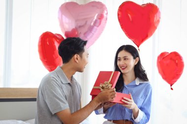 Mutlu Asyalı çift birlikte aşklarını kutluyor, erkek arkadaş sürpriz sevgiliye kırmızı hediye kutusu veriyor, romantik aşık sevgililer gününü güzel dekore edilmiş yatak odasında kutluyor.