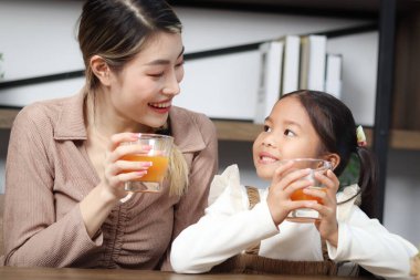Evde oturma odasında otururken taze portakal suyu içen Asyalı anne ve kız çocuk sağlıklı beslenmek için çocuğa bakıyor. Mutlu aile birlikte vakit geçiriyor..