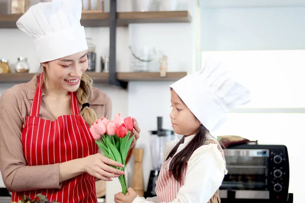 Happy Asian mother and daughter wear apron chef hat at kitchen, girl gives beautiful tulip flowers for mom during prepare meal together. Cute kid chef and family cooking food at home. Happy mother day