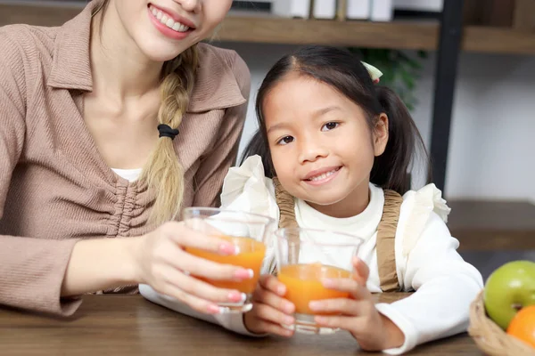 Portrait of Asian daughter girl child drinking fresh orange juice with mother during sitting in living room at home, parent take care kid to get healthy diet. Happy family spending time together.