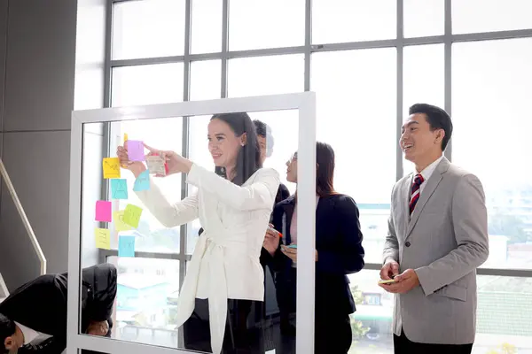 Businesspeople brainstorming at the meeting. Businessmen and businesswomen discussing, writing and putting paper note on summary chart glass board for creative idea, critical thinking teamwork,