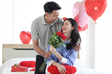 Mutlu Asyalı adam sevgilisine güzel kırmızı gül demeti verirken beyaz yatakta oturan romantik çift Sevgililer Günü 'nde birlikte unutulmaz bir yıldönümü kutluyor..