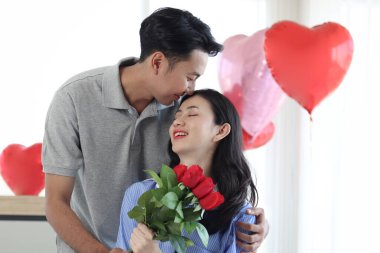 Mutlu Asyalı adam sevgilisine güzel kırmızı gül demeti veriyor ve beyaz yatakta otururken öpüşüyor. Romantik çift sevgililer gününde birlikte unutulmaz bir yıldönümü kutluyor.