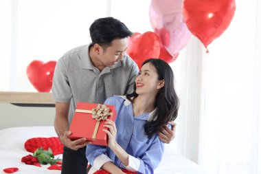 Mutlu Asyalı adam yatak odasında beyaz yatakta otururken sevgilisine kırmızı güzel bir hediye kutusu veriyor. Romantik çift Sevgililer Günü 'nde birlikte unutulmaz bir yıldönümü kutluyor..